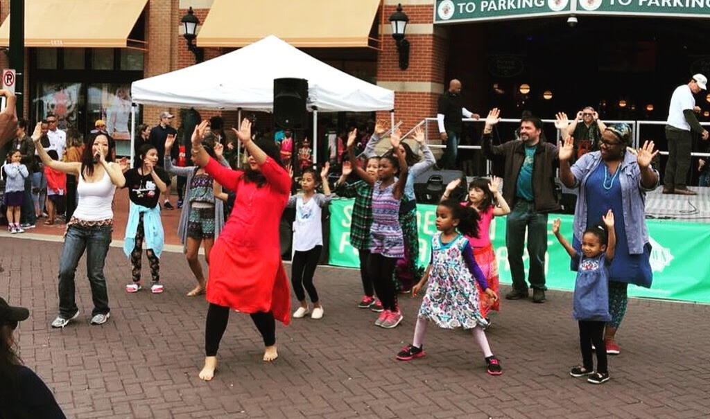 A group of kids and adults dances outside, hands raised